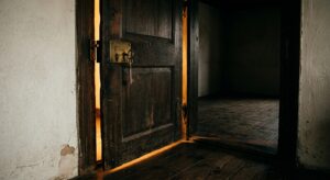 Heavy old wooden door slightly ajar with a brass lock and key, warm light pouring through the gap onto dark floorboards, an empty bare room visible beyond